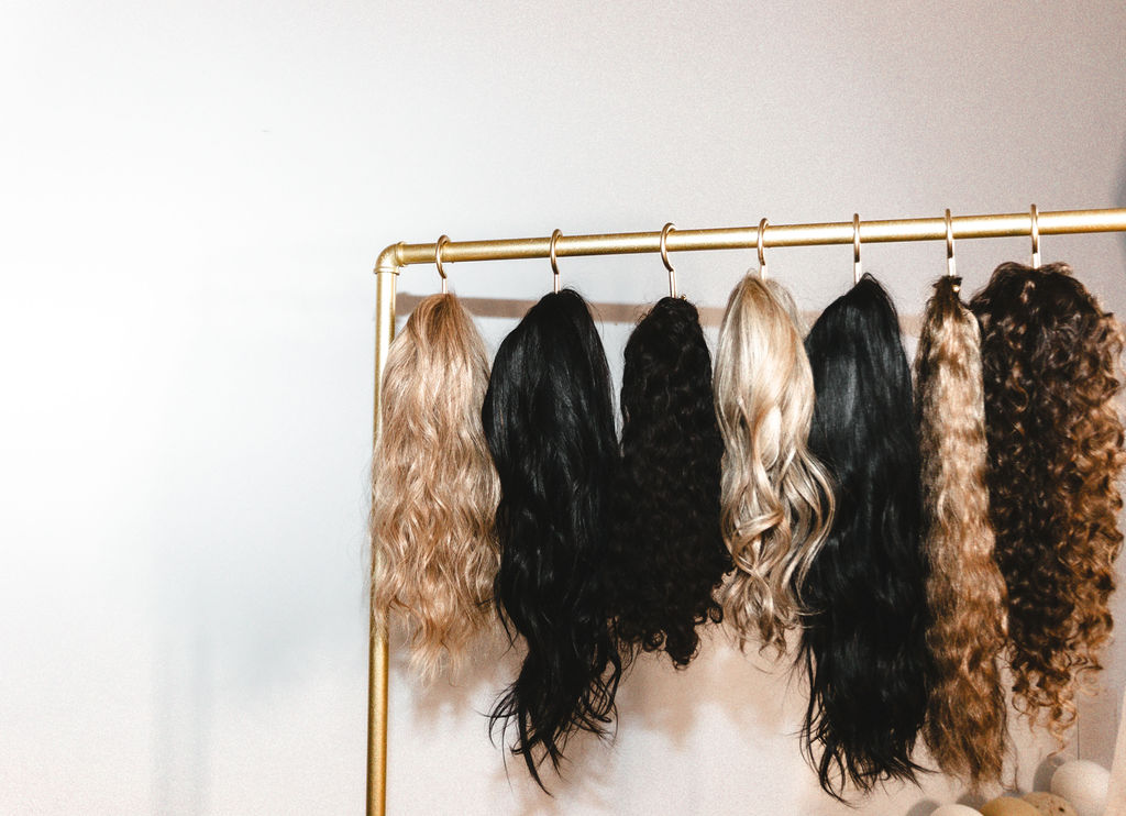 A diverse selection of hair extensions in various textures and shades, from blonde waves to deep black curls, displayed on a gold rack at Garnish Collective.